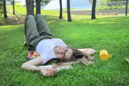 Téléchargez les photos : Heureuse étudiante se reposer dans un parc sur une herbe, en regardant la caméra - en image libre de droit