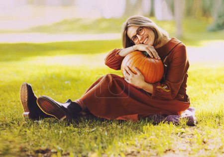 Téléchargez les photos : Belle femme ont un loisir posant dans le parc d'automne habillé en robe brune longue, fille posant avec citrouille - en image libre de droit