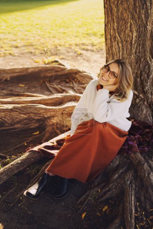 Foto de Hermosa mujer joven tienen un ocio sentado en el parque de otoño bajo el árbol con enormes raíces, vestido con falda larga marrón y suéter blanco - Imagen libre de derechos