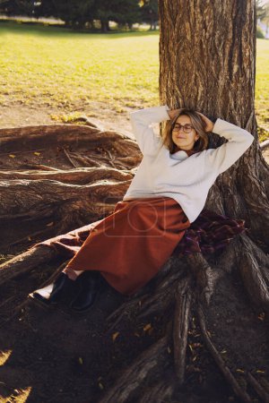 Foto de Hermosa mujer joven tienen un ocio durmiendo en el parque de otoño vestida con falda larga marrón y suéter blanco - Imagen libre de derechos
