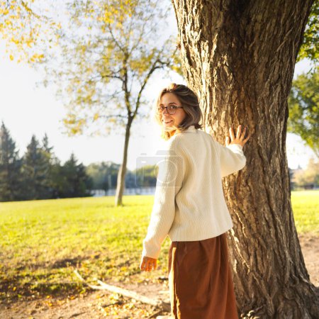 Foto de Hermoso retrato de mujer joven sonriente en el parque de otoño vestido con falda larga marrón y suéter blanco - Imagen libre de derechos