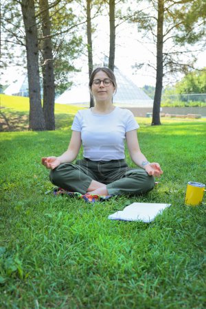 Téléchargez les photos : Heureuse étudiante se reposer en faisant du yoga dans un parc avec un livre sur une herbe, en regardant la caméra - en image libre de droit