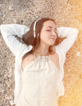 Téléchargez les photos : Femme souriante couchée sur un sable marin et écoutant de la musique par des écouteurs, ferma les yeux - en image libre de droit