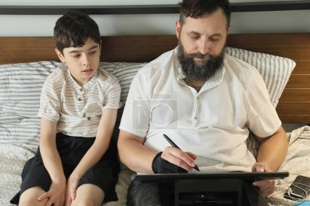 Téléchargez les photos : Graphiste à distance travaillant à la maison sur un lit, son fils assis près de lui - en image libre de droit