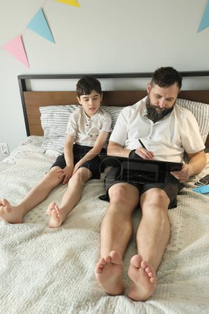 Téléchargez les photos : Graphiste à distance travaillant à la maison sur un lit, son fils assis près de lui - en image libre de droit