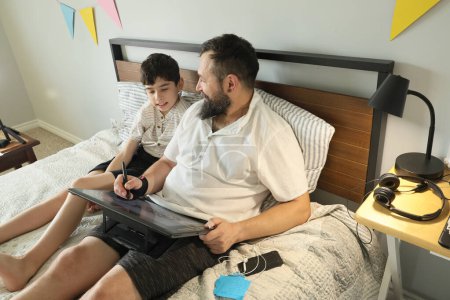 Téléchargez les photos : Graphiste à distance travaillant à la maison sur un lit, son fils assis à proximité - en image libre de droit