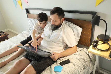 Téléchargez les photos : Graphiste à distance travaillant à la maison sur un lit, son fils l'aidant - en image libre de droit