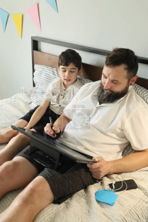 Téléchargez les photos : Graphiste homme à distance travaillant à la maison sur un lit, son fils assis près de lui - en image libre de droit