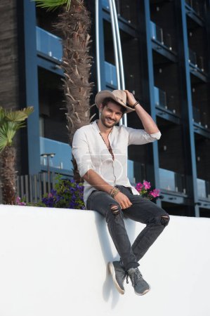 Foto de Hombre guapo retrato de moda vestido con sombrero de paja y camisa blanca, foto al aire libre contra el paisaje turístico de la ciudad tropical - Imagen libre de derechos