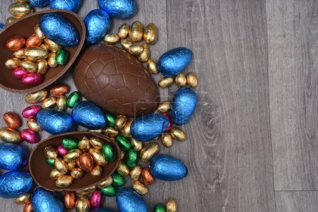 Pile or group of multi colored and different sizes of colourful foil wrapped chocolate easter eggs in pink, blue, gold and green. Large halves of a brown milk chocolate egg have mini eggs inside, on a grey wooden background. 