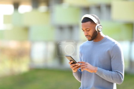 Téléchargez les photos : Homme noir marchant vérifier la musique sur le téléphone intelligent dans la rue - en image libre de droit