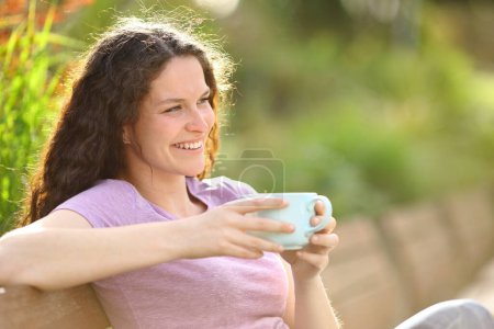 Photo for Happy woman drinking coffee smiling sitting on a baench in a park - Royalty Free Image