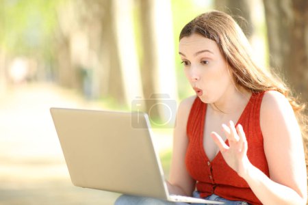 Foto de Mujer sorprendida revisando el contenido del portátil sentado solo en un parque - Imagen libre de derechos