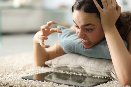 Téléchargez les photos : Femme en colère vérifier le contenu de la tablette couché sur le sol à la maison - en image libre de droit