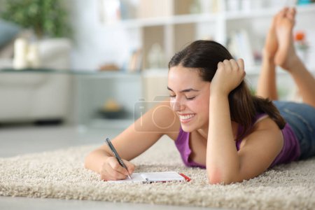 Téléchargez les photos : Femme heureuse prenant des notes dans l'ordre du jour couché sur un tapis à la maison - en image libre de droit