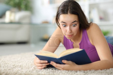 Téléchargez les photos : Surprise femme lisant un livre de papier couché sur un tapis à la maison - en image libre de droit