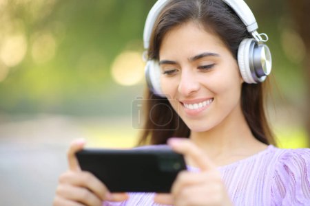 Glückliche Frau schaut sich Video am Telefon mit Kopfhörer in einem Park an