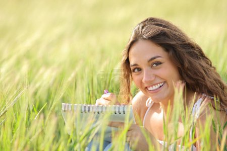 Glückliche Frau zeichnet auf einem Notizbuch in einem Feld, das dich ansieht