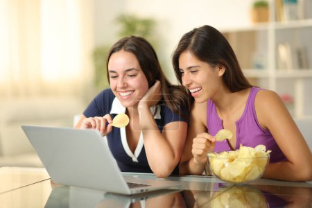 Two happy friends watching movie on laptop eating snack sitting at home