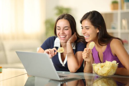 Las mujeres felices comiendo papas fritas y viendo vídeo en el ordenador portátil en casa