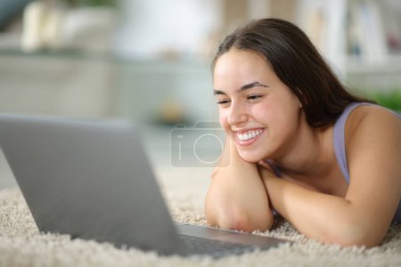 Mujer feliz acostada en el suelo viendo los medios de comunicación en el ordenador portátil en casa