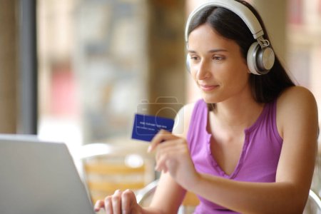 Frau kauft mit Laptop und Kreditkarte online und trägt Kopfhörer auf der Terrasse eines Cafés
