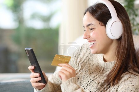 Mujer feliz con auriculares medios de compra en línea con tarjeta de crédito y teléfono inteligente en casa