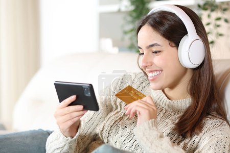 Photo for Happy woman wearing headphone buying media products with credit card and phone at home - Royalty Free Image