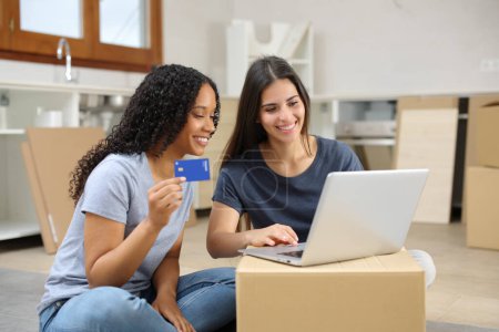 Two happy roommates moving home and buying online with laptop and credit card in the kitchen