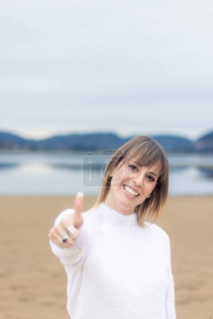 Téléchargez les photos : Beauté rayonnante sur la plage : Femme souriante dans un pull blanc donnant un pouce - en image libre de droit