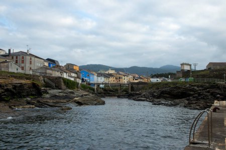 serene coastal town with colorful houses, calm waters, rocky shores, under a cloudy sky, surrounded by distant green hills