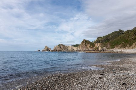 tranquil pebble beach with clear waters, rocky formations, and lush greenery under a partly cloudy sky