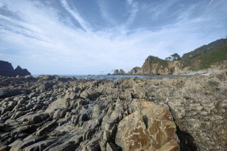 rugged shoreline with jagged rock formations, calm sea waters, and a clear sky with wispy clouds. It's a serene and natural setting