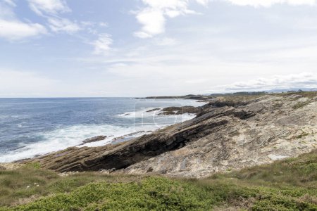rocky coastline with greenery, overlooking a vast, serene ocean under a partly cloudy sky.