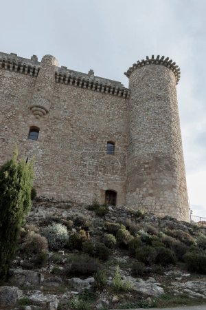 Téléchargez les photos : Un château historique en pierre avec des tours cylindriques, des murs crénelés et de petites fenêtres, situé dans un paysage rocheux et végétalisé - en image libre de droit