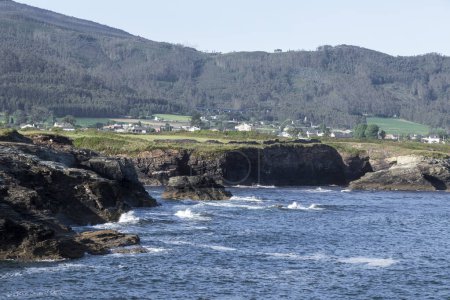 coastal landscape with rocky shores, turbulent waters, and a green, hilly backdrop featuring a distant village