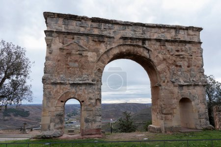 ancient, weathered stone arch with two openings stands against a backdrop of a cloudy sky and distant landscape