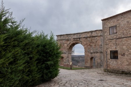 old stone building with a large archway, adjacent to a green bush, under a cloudy sky