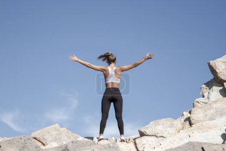 A person stands triumphantly atop rocky terrain, arms outstretched, under a clear blue sky, embodying freedom and achievement