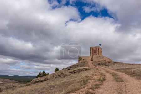 old stone structure on a hill, path leading up, flag on top, surrounded by a rugged, cloud-shadowed landscape