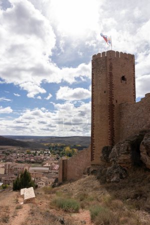 old stone tower with a flag, overlooking a scenic town amidst nature, under a cloudy sky