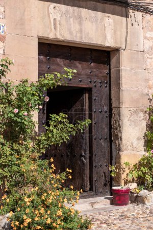 Verwitterte Holztür mit blühenden Blumen und einem roten Eimer in einer Steinmauer.