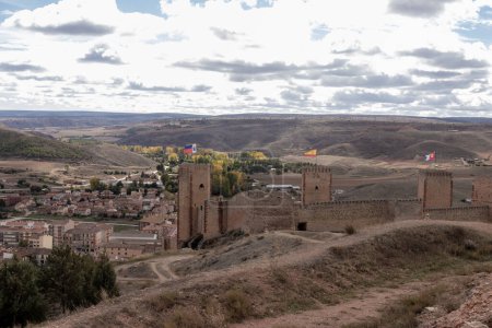 Téléchargez les photos : Ancien château en pierre avec trois drapeaux, surplombant une ville dans une vallée au milieu de collines et d'un ciel nuageux - en image libre de droit