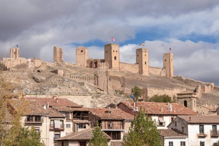 castillo de piedra, pueblo rústico debajo, banderas en las torres, cielo parcialmente nublado, ambiente histórico, luz de la mañana o de la tarde
