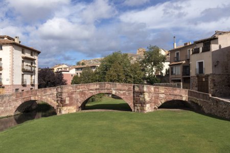stone bridge spans a green area, connecting quaint houses under a partly cloudy sky in a historic setting