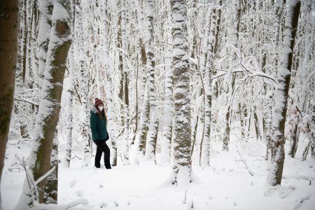 Eine einsame Gestalt steht in einem schneebedeckten Wald, in den Hintergrund ragen weiß verhüllte Bäume. Die Person trägt eine Jacke in Krickente und eine Mütze, und eine weiße Maske bedeckt ihr Gesicht, was die Einsamkeit der Winterkulisse unterstreicht.