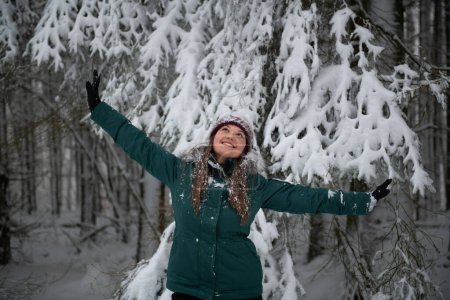 The image depicts a woman in a snow-filled forest, arms joyfully raised. She is wearing a teal winter jacket and a snow-speckled knitted hat, with a backdrop of heavily snow-laden branches