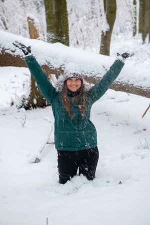Téléchargez les photos : Une femme vêtue d'un manteau d'hiver sarcelle les bras levés joyeusement au milieu d'une forêt enneigée. Elle est profondément genou dans la neige, et son chapeau est légèrement saupoudré de flocons de neige, améliorant l'atmosphère hivernale. - en image libre de droit