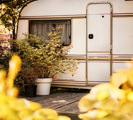 Weiße Wohnwagenanhänger auf einer grünen Wiese zwischen Büschen und Bäumen auf einem Campingplatz. Sommerlandschaft. Europa. Lebensstil von Menschen, die Reisen, Tourismus, Wohnmobil und Wohnmobil lieben.