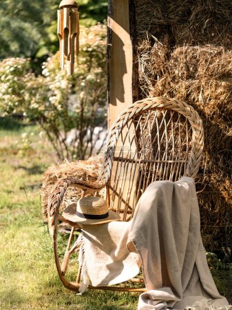 Photo for Wicker chair with a plaid and a straw hat in the garden. Armchair against the background of a straw house. The state of rest and tranquility. Singing winds on the house. - Royalty Free Image